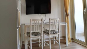 Ein Zimmer mit an der Wand montiertem Fernseher, einem Schreibtisch mit zwei Stühlen und einer Glastür mit Vorhängen.