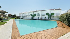Et rektangulært swimmingpool med en træterrasse, omgivet af en række hvide bygninger med altaner og grønt.
