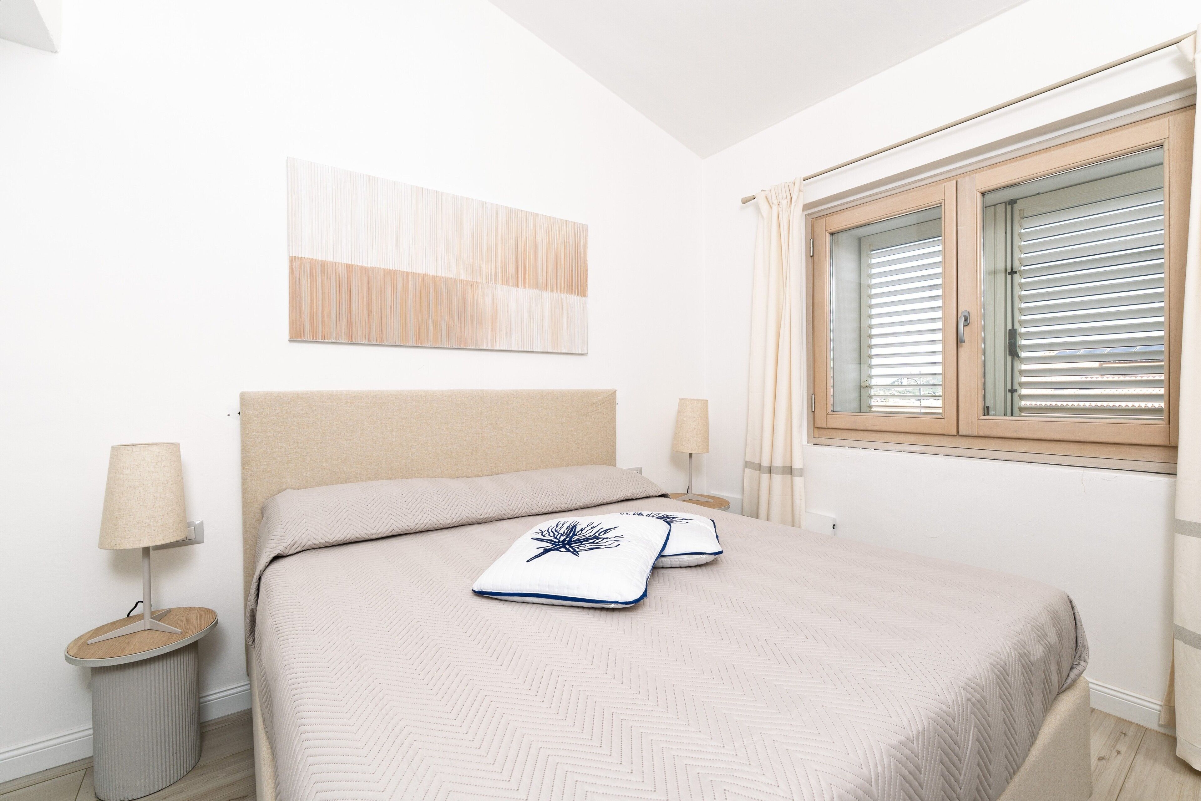 Un dormitorio con cama, dos lámparas de noche, una mesita de noche, una ventana con persianas y una obra de arte colgada en la pared.