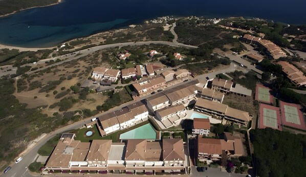 Vista aérea de un complejo turístico frente al mar, con varias edificaciones, una piscina y un camino que conduce a la playa.