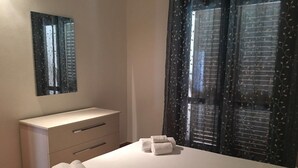 Een slaapkamer met een bed, nachtkastje met spiegel, kapstok en raam met gordijnen.