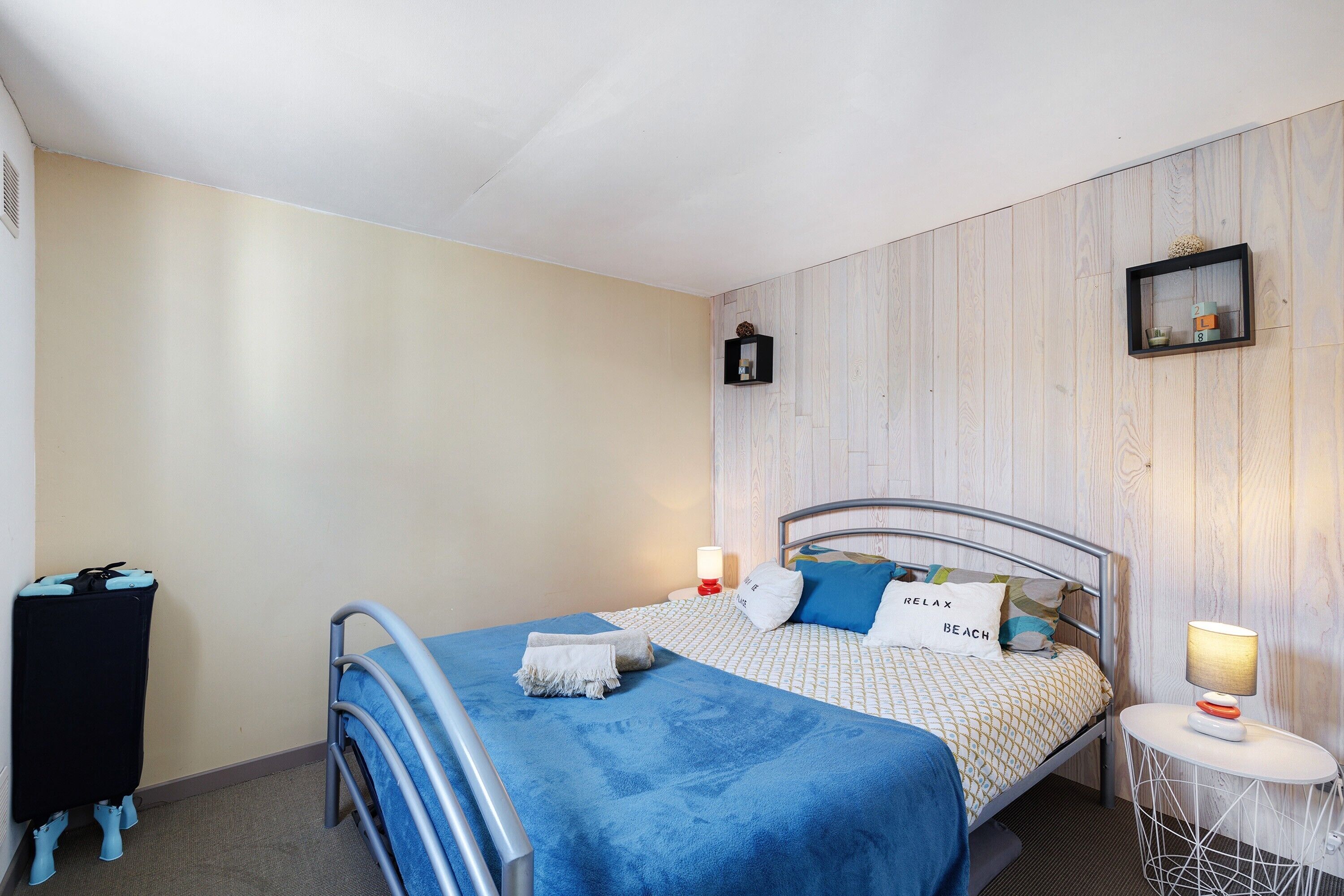 Un lit simple avec une couverture bleue, une table de chevet avec une lampe, une petite étagère avec des objets décoratifs et un cadre photo fixé au mur.