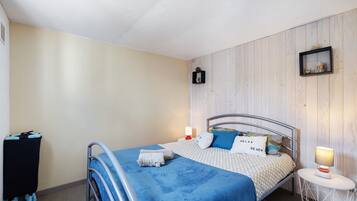 Un lit simple avec une couverture bleue, une table de chevet avec une lampe, une petite étagère avec des objets décoratifs et un cadre photo fixé au mur.