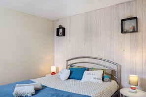 A neatly made bed with a patterned bedspread, pillows with text, a bedside table with a lamp, and a wall-mounted shelf with decorative items. - Holiday Home “La Petite Vague” with Private Terrace and Garden (Saint-Hilaire-de-Riez)
