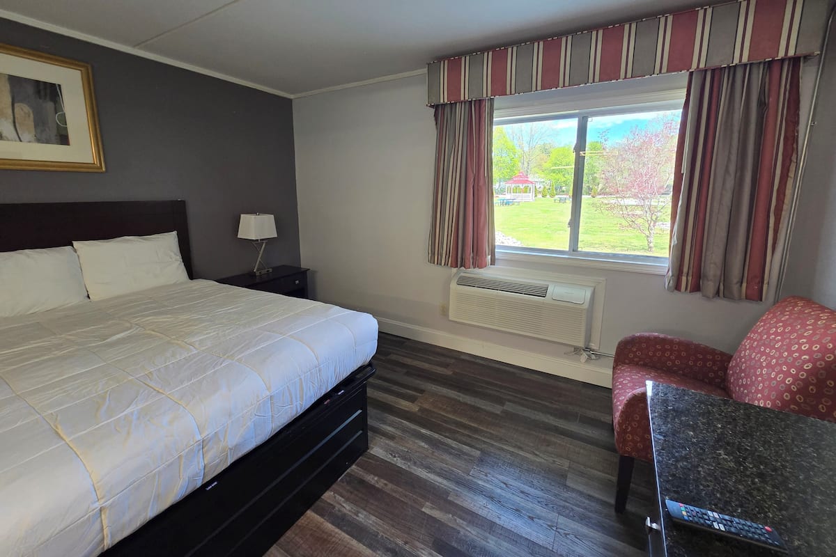 A hotel room with a bed, a nightstand, a lamp, a window with curtains, a radiator, and a small table with a chair.