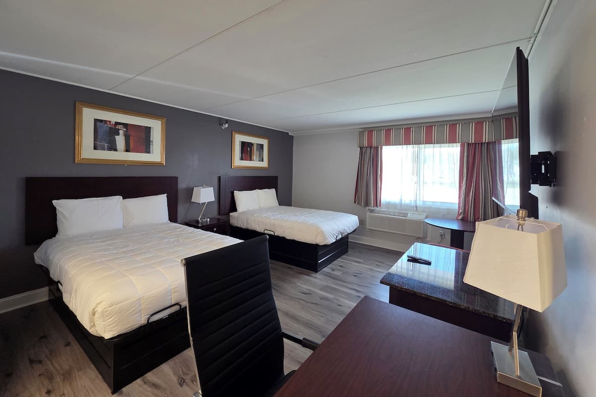 A hotel room with two beds, a desk, a chair, a lamp, a window with curtains, and framed pictures on the wall.
