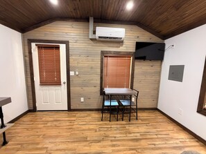 A room with wooden flooring, two doors with blinds, a table with chairs, and an air conditioning unit on the wall.