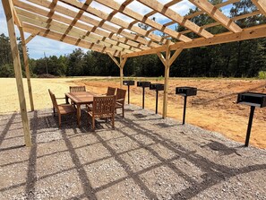 A wooden pergola structure with a table and chairs underneath, surrounded by a gravel area with mailboxes.