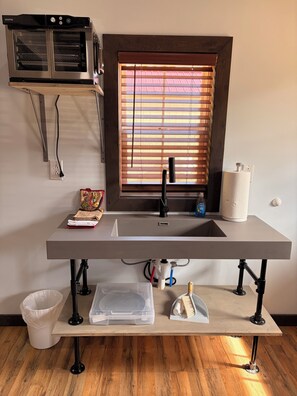 A kitchen with a built-in oven, a window with blinds, a sink, and a countertop with various items.