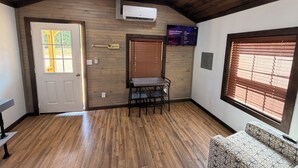 Una habitación con piso de madera, una puerta con ventana, una mesa con sillas, un aire acondicionado instalado en la pared y un televisor de pared mostrando un menú.