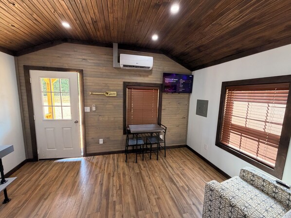 A room with wooden flooring, a door with a window, a table with chairs, a mounted air conditioner, and a wall-mounted TV displaying a menu.