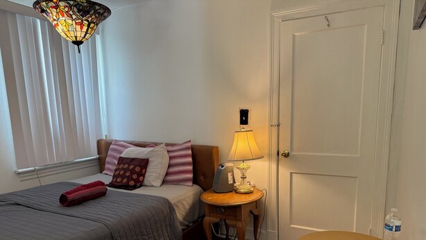 Ein Schlafzimmer mit Bett, Nachttisch, Lampe, Sessel und einer Tür.