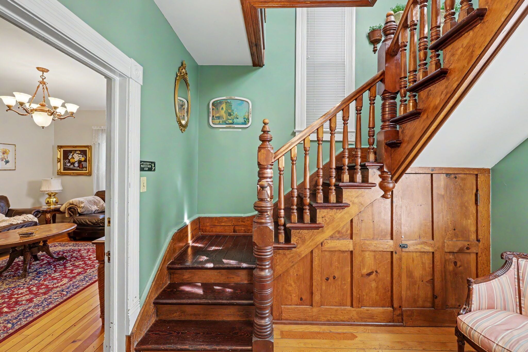 A staircase with wooden balusters and a handrail, a chandelier, a framed picture, and a small table with a rug.