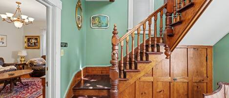 A staircase with wooden balusters and a handrail, a chandelier, a framed picture, and a small table with a rug.