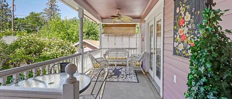 A covered porch with a ceiling fan, white wicker chairs, a glass table, and a floral painting on the wall.