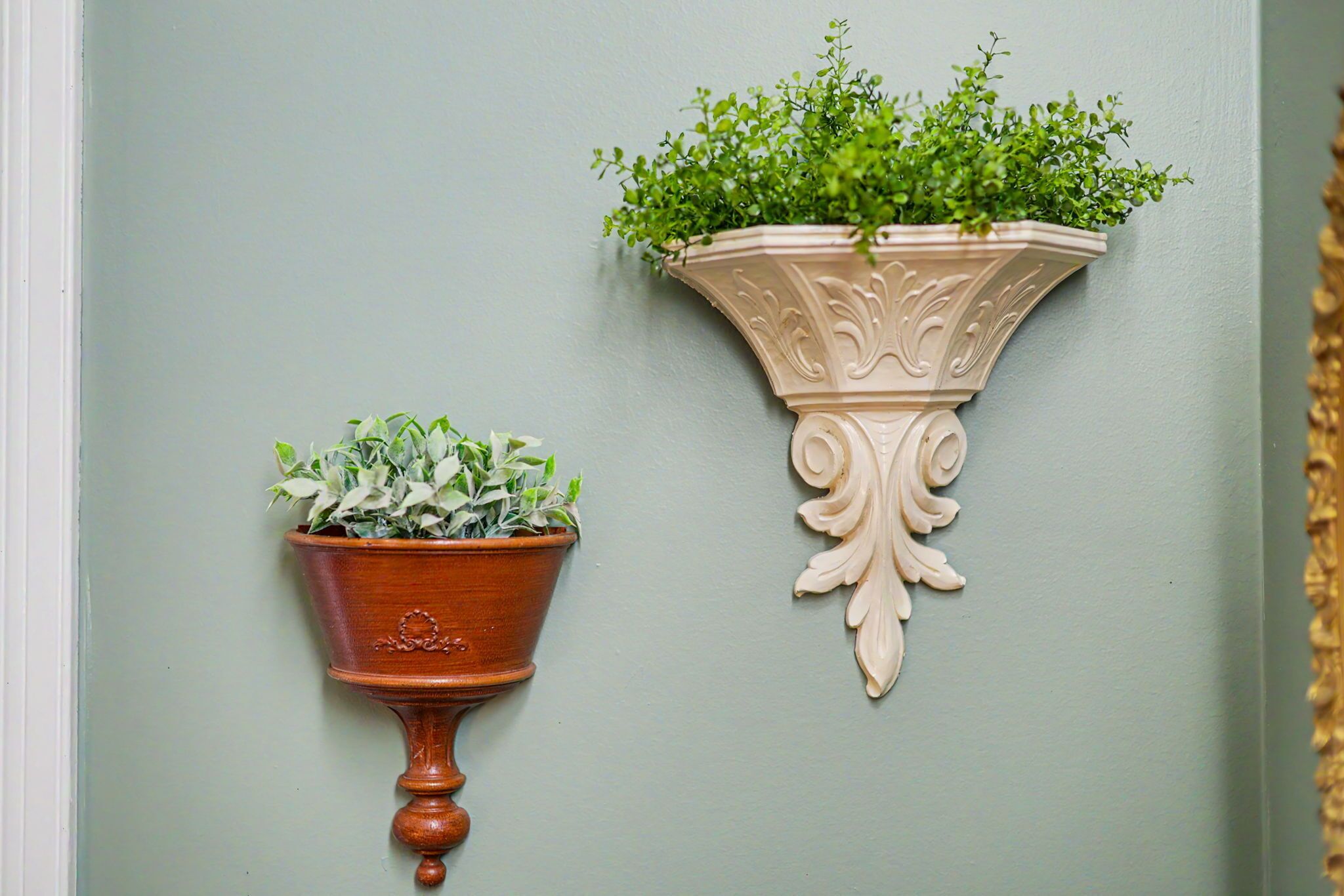 Two wall-mounted planters with plants, one in a brown pot and the other in a cream pot, against a light green wall.