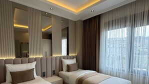 A modern hotel room with a large bed, a smaller bed, a nightstand, a mirror, and a view of the city. - Luxury apartment in Alanya with 270° panoramic views & only 5 min to the beach (Kargicak)