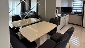 A modern kitchen with a dining area, featuring a central table with chairs, a built-in oven, and a window with blinds. - Luxury apartment in Alanya with 270° panoramic views & only 5 min to the beach (Kargicak)