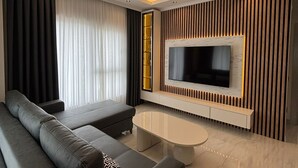 A modern living room with a large flat-screen TV, a dark grey sofa, a small coffee table, and a wooden accent wall. - Luxury apartment in Alanya with 270° panoramic views & only 5 min to the beach (Kargicak)