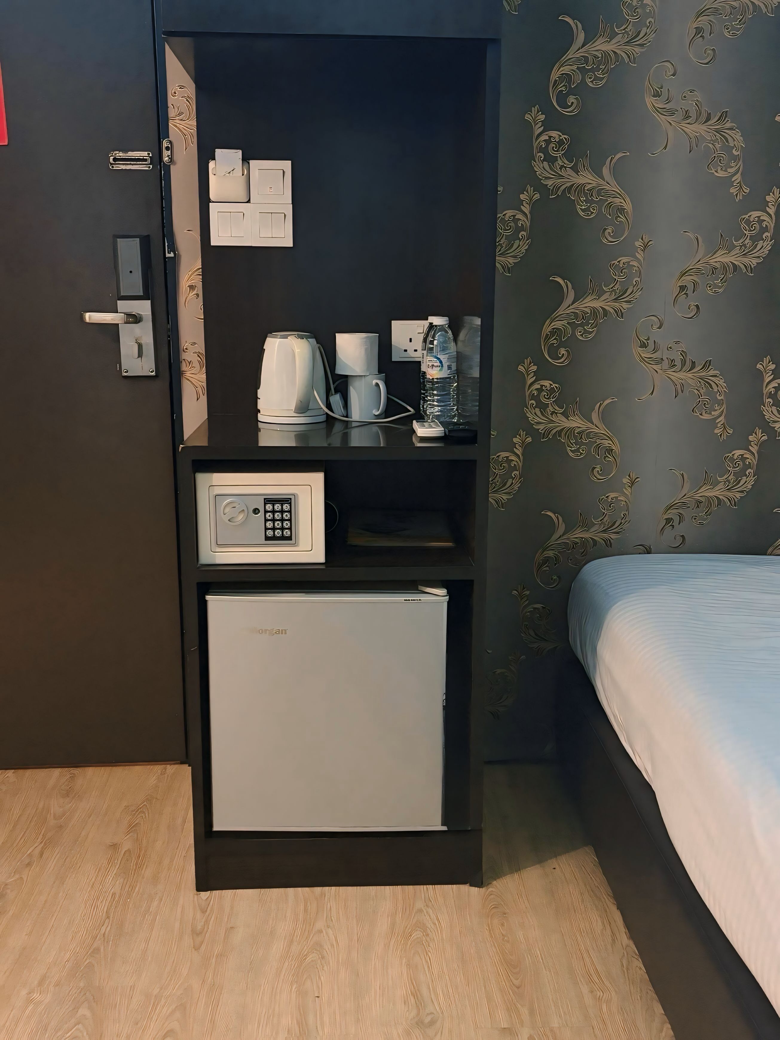 A compact room with a bed, a small refrigerator, a coffee maker, a water bottle, and a power outlet.