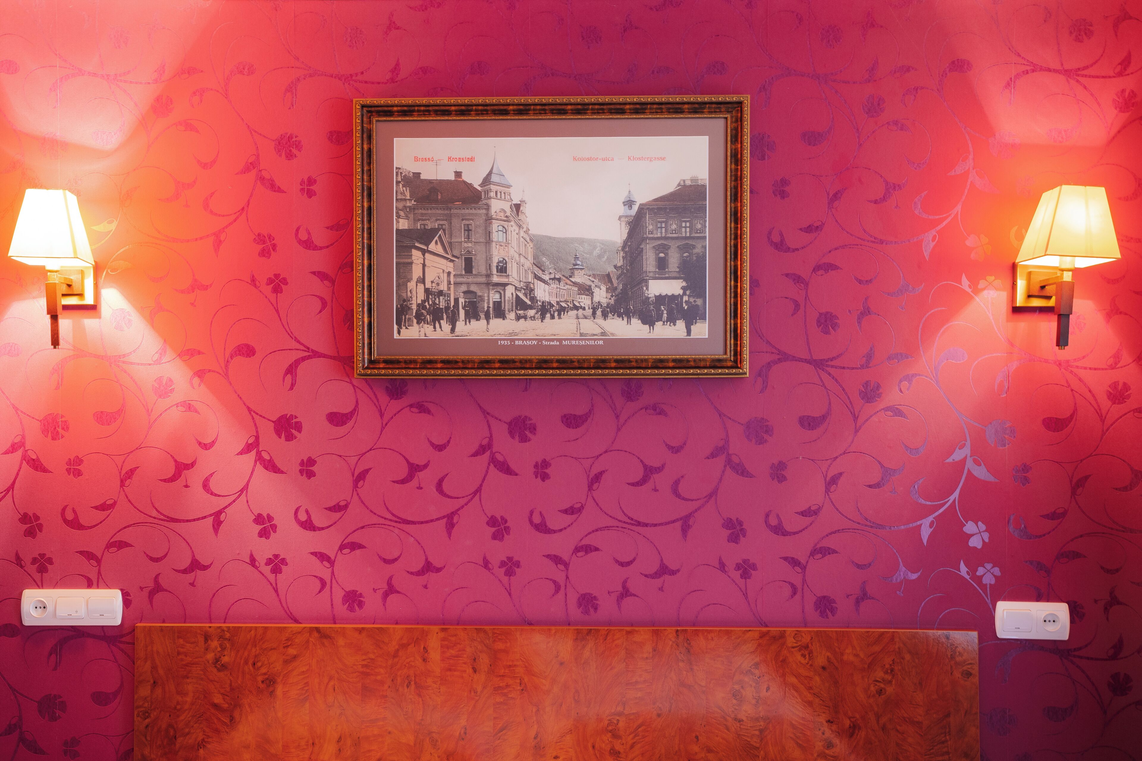 A framed historical photograph on a wall with patterned wallpaper, two wall-mounted lamps, and a wooden panel below.