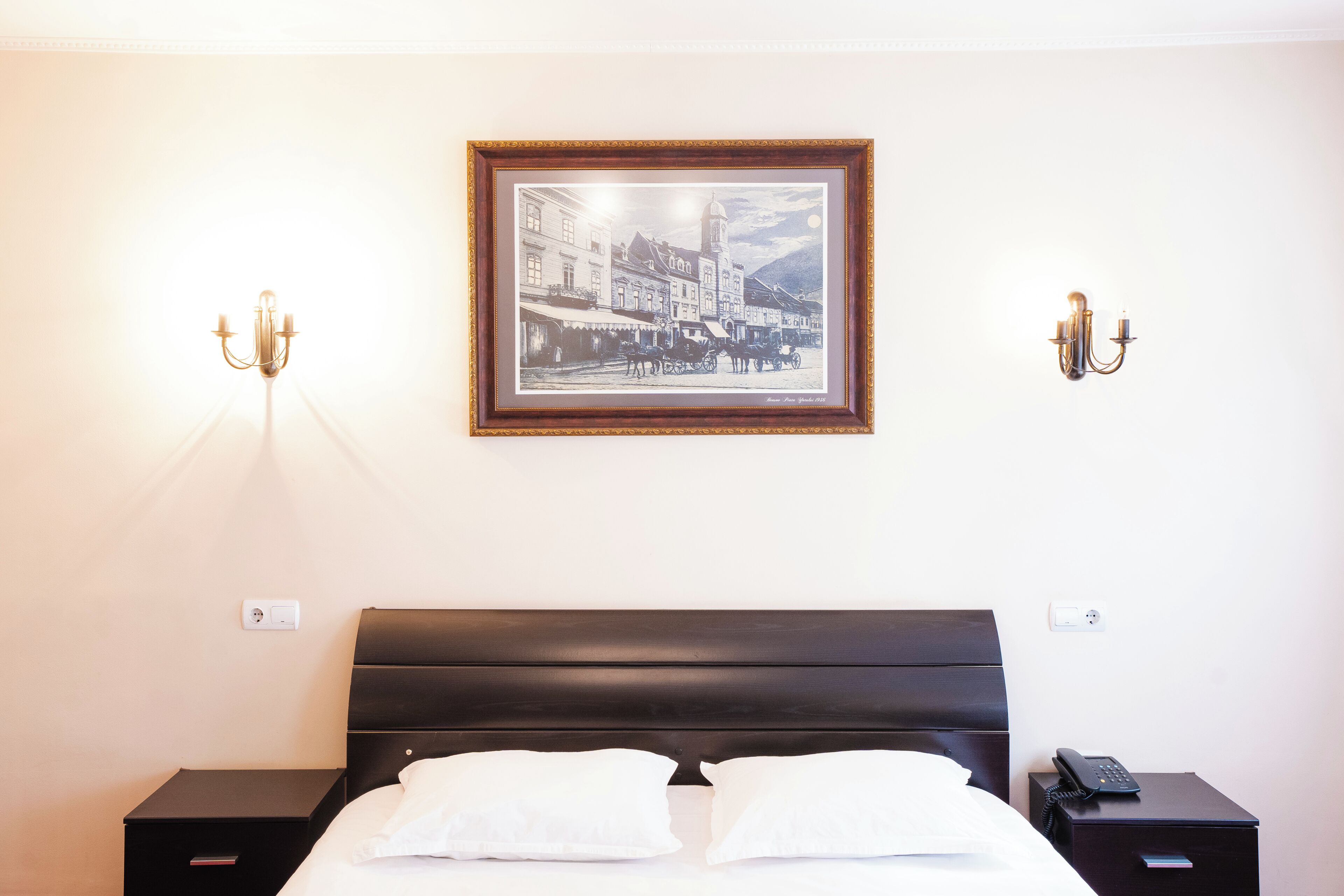 A hotel room with a bed, a framed picture on the wall, two bedside lamps, and a nightstand with a phone.