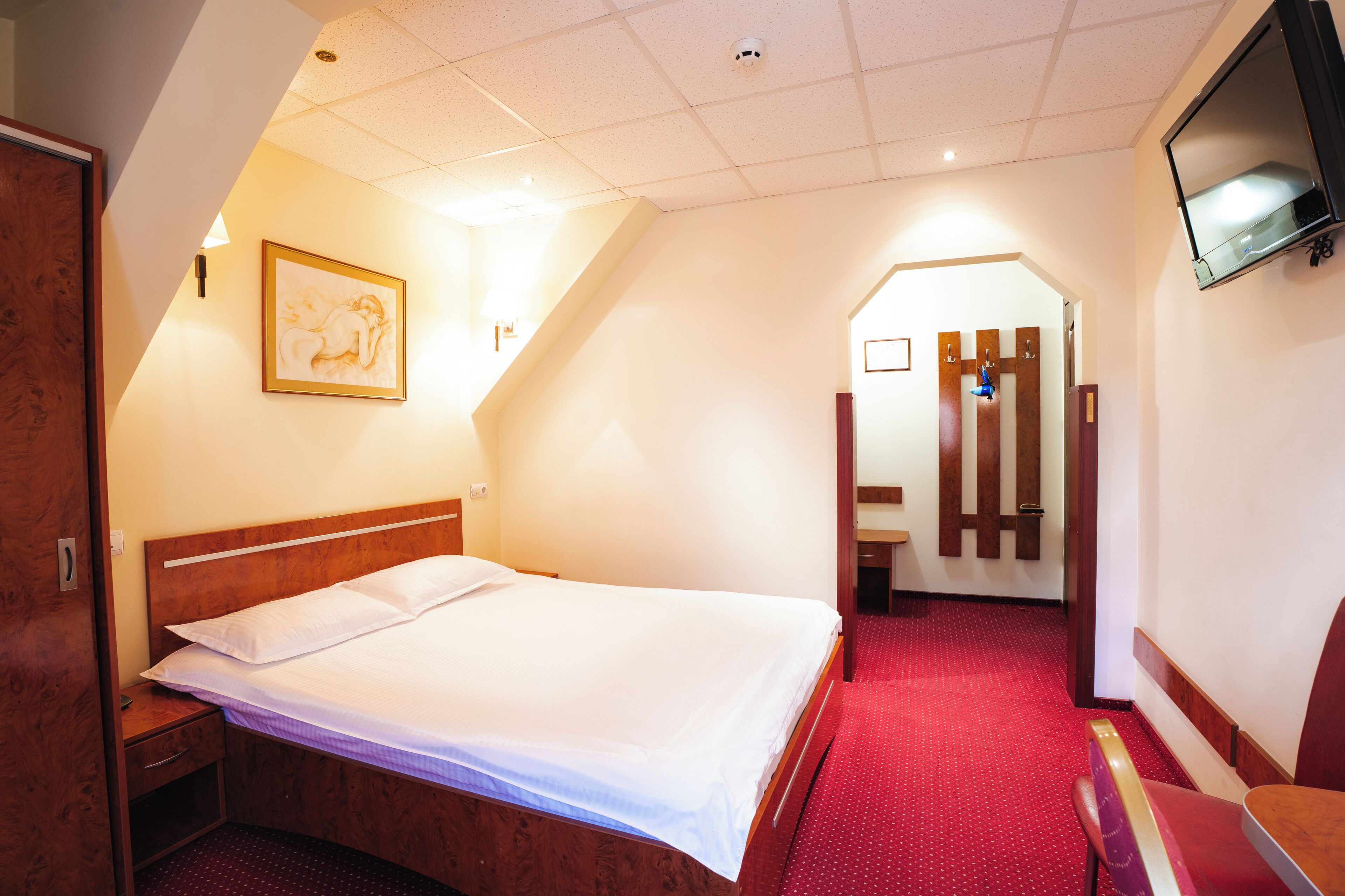 Habitación de hotel con cama, mesita de noche de madera, televisor montado en la pared y suelo cubierto de alfombra roja.