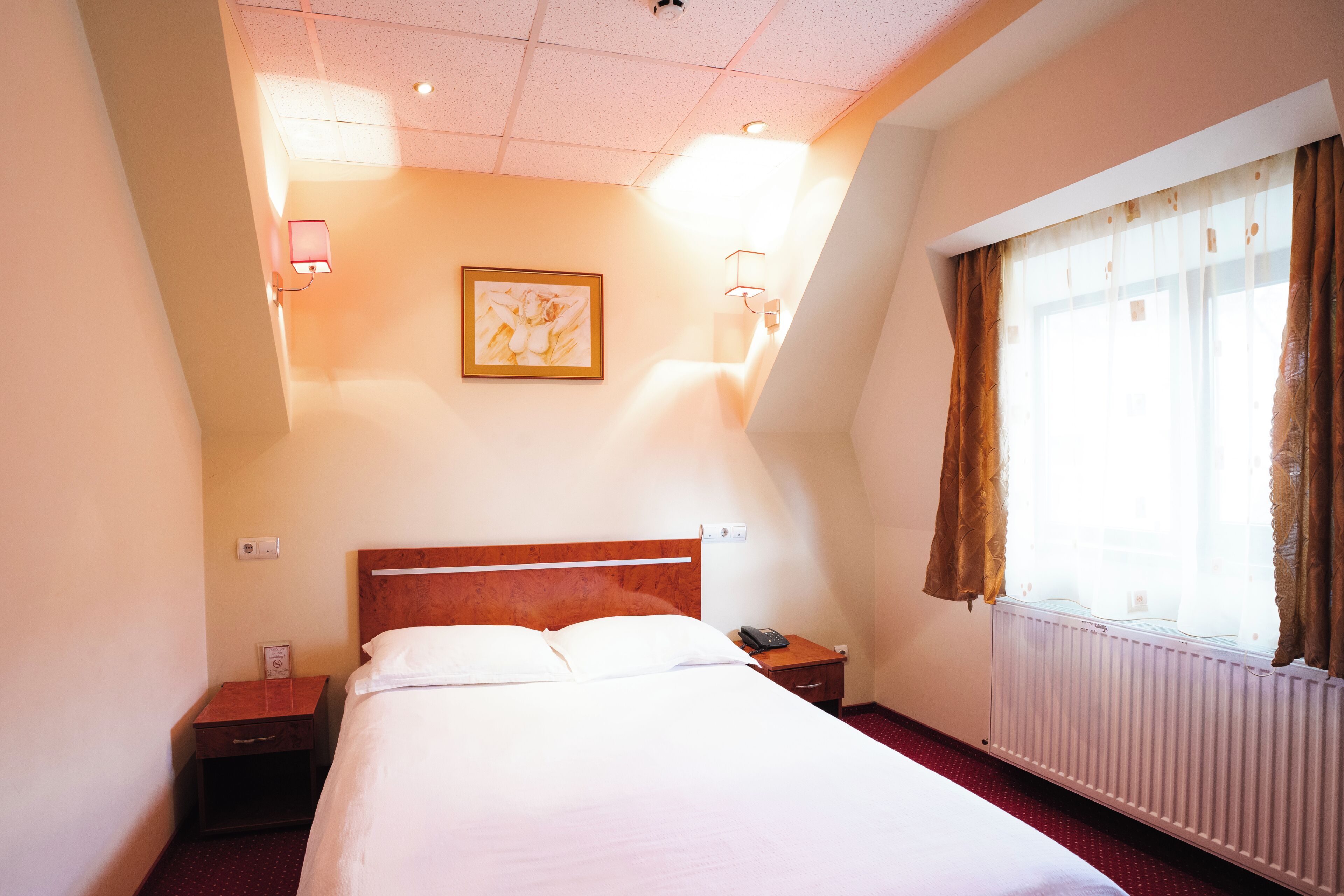 Una habitación de hotel con cama, mesita de noche, lámpara de pared y ventana con cortinas.