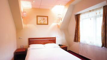 Una habitación de hotel con cama, mesita de noche, lámpara de pared y ventana con cortinas.