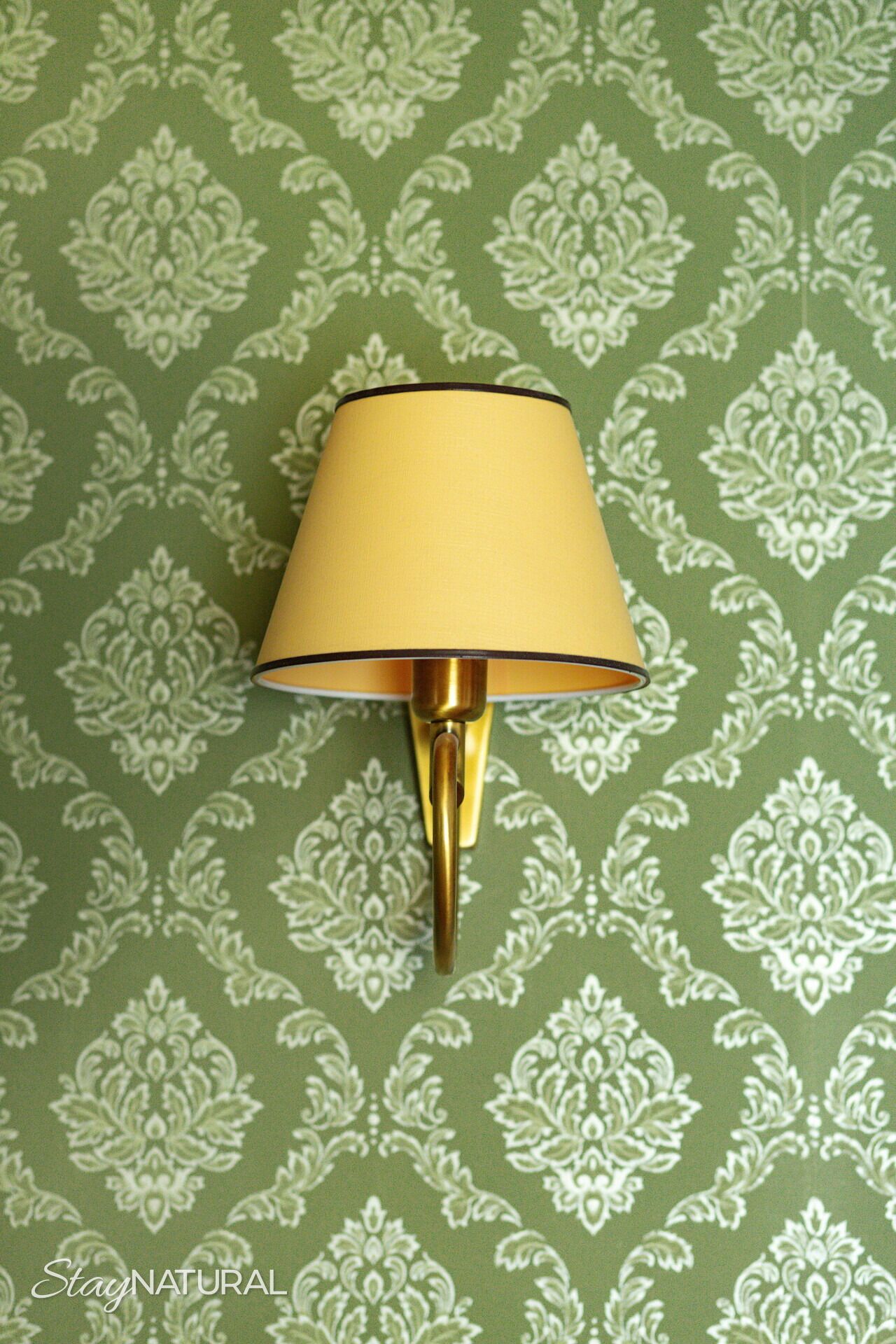 A wall-mounted lamp with a yellow shade against a green patterned wallpaper.