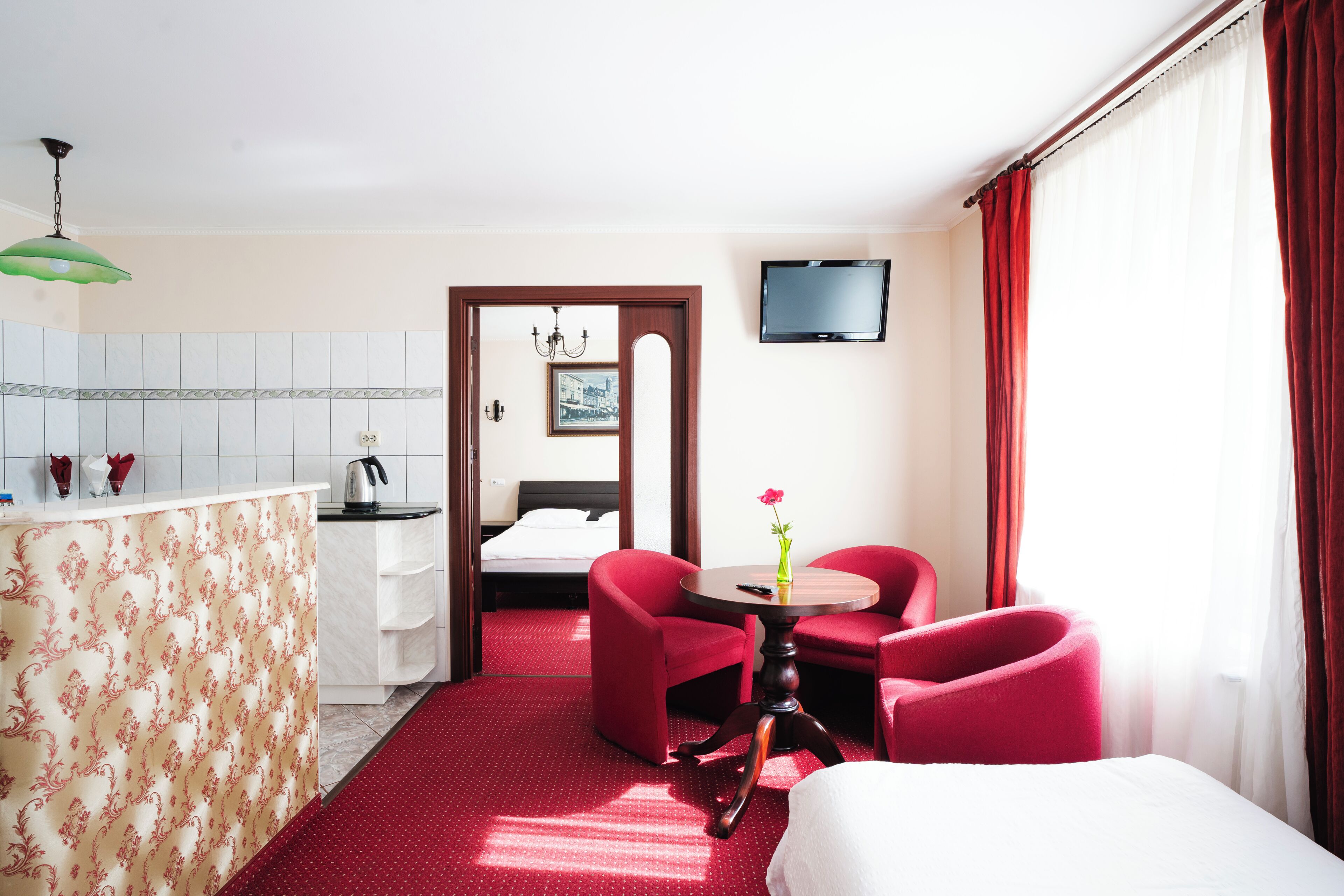 A hotel room with a bed, a desk with a chair, a small table with a vase, and a TV on the wall.