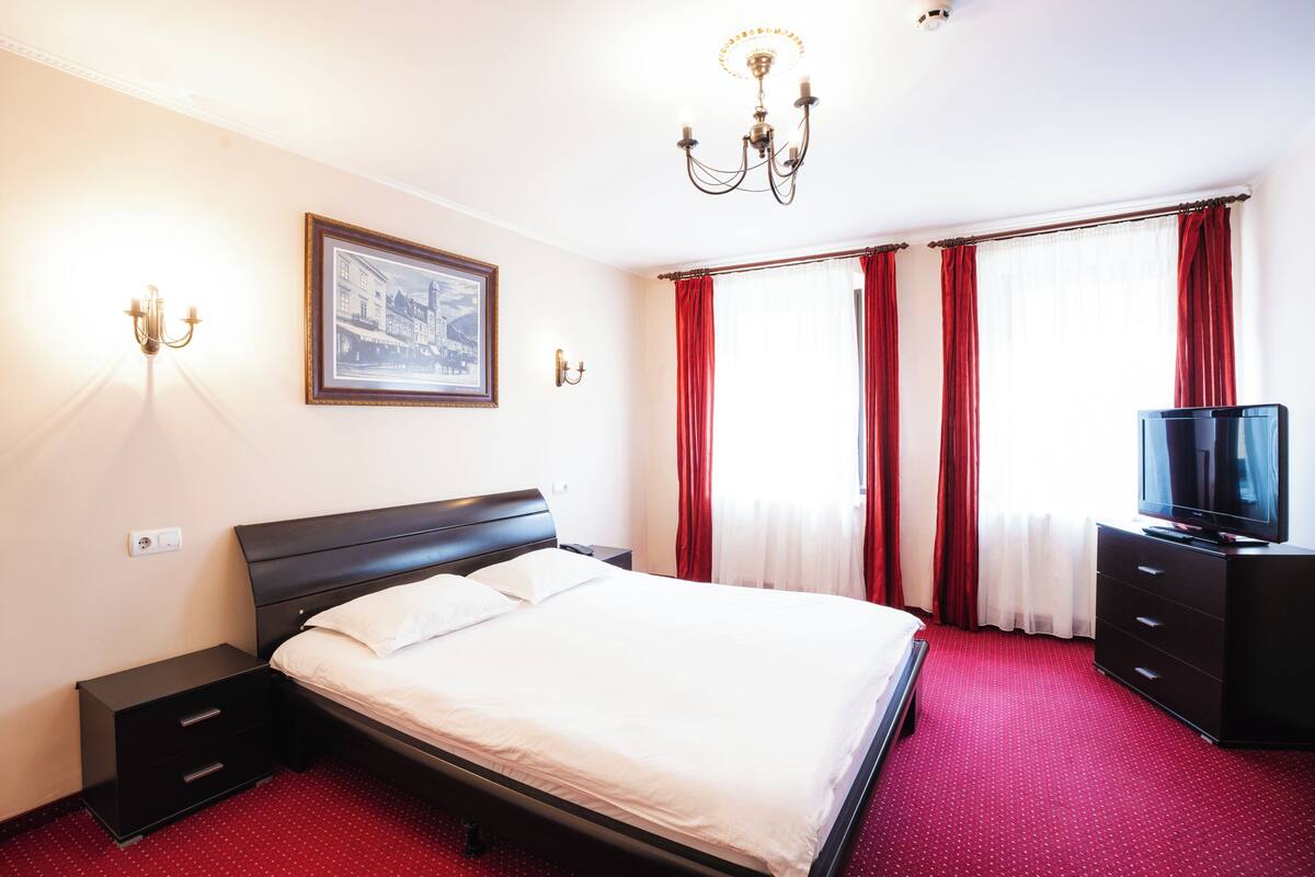 Habitación de hotel con cama, televisor, mesita de noche, cómoda, candelabro y ventana con cortinas.