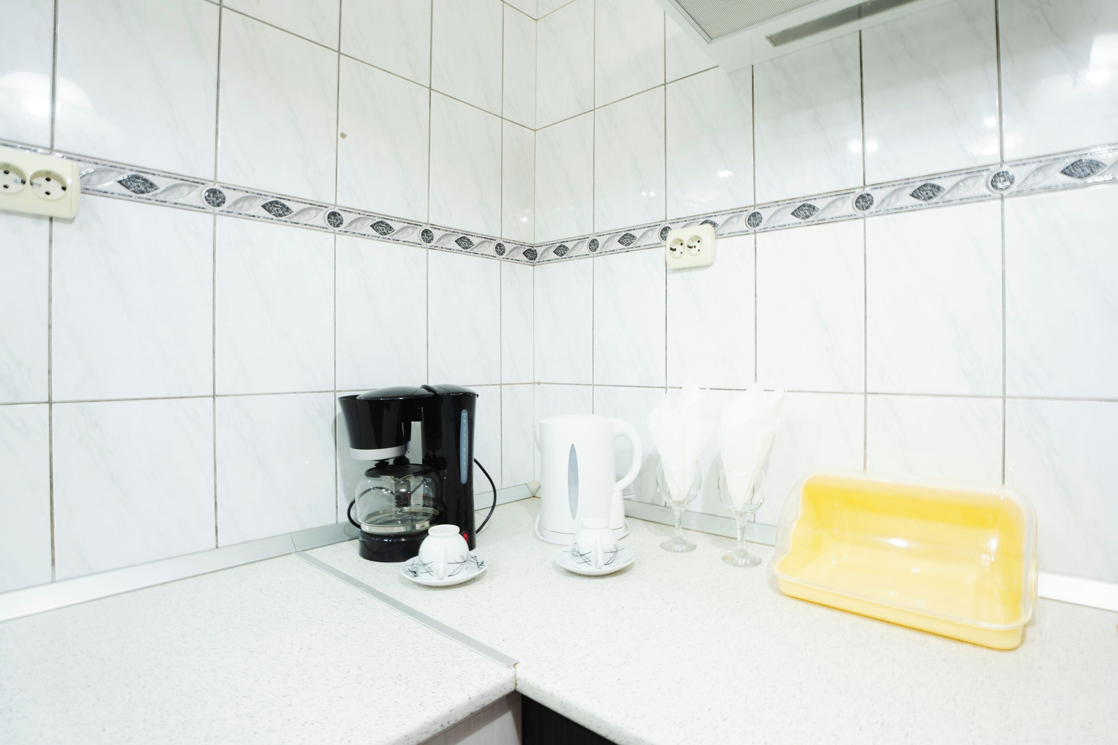 Une cuisine avec des carreaux blancs, une machine à café, une tasse blanche, un égouttoir jaune et une prise de courant.