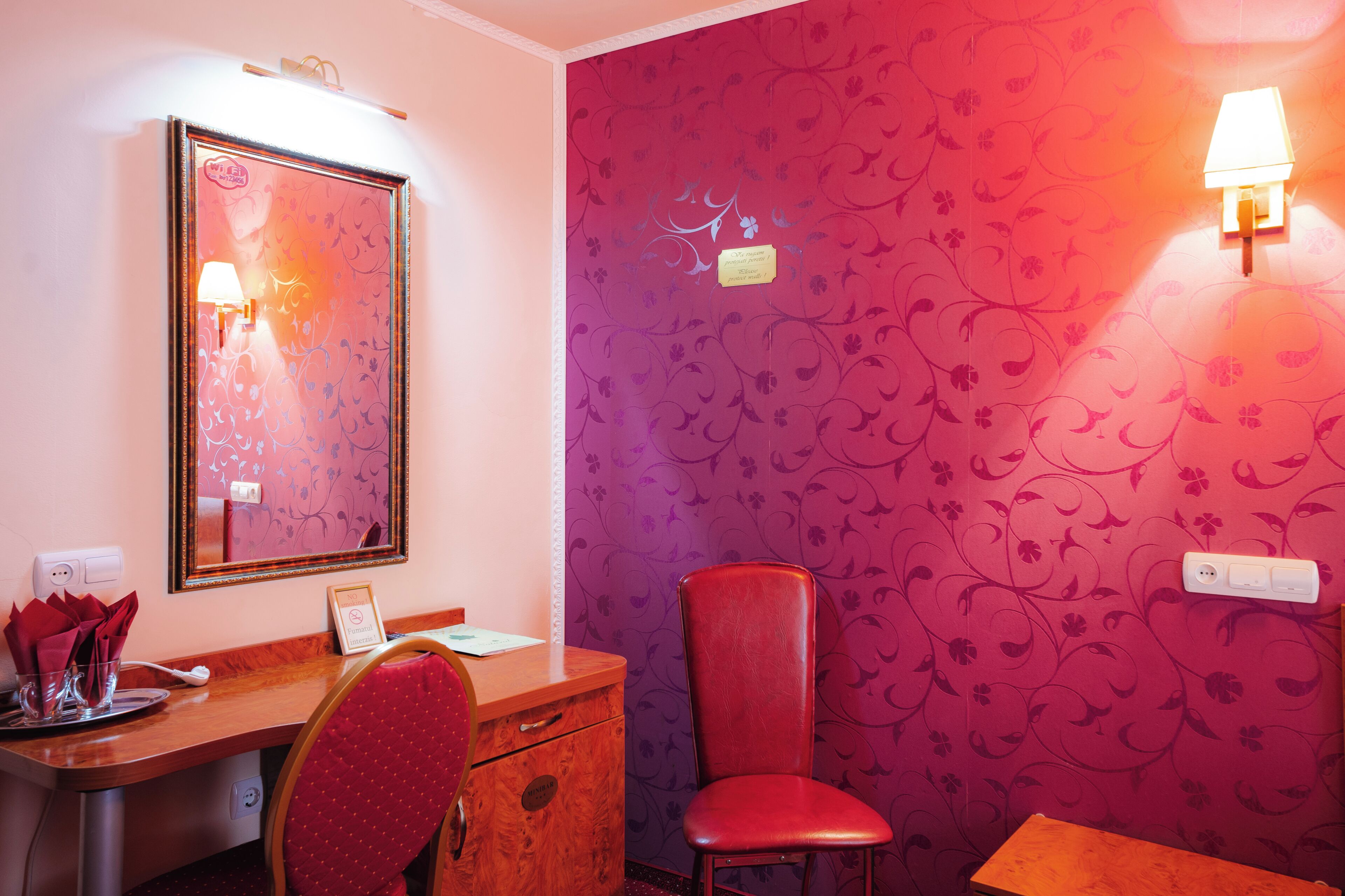 Une pièce avec un bureau en bois, une chaise rouge, un miroir et un mur recouvert d’un papier peint à motifs floraux.