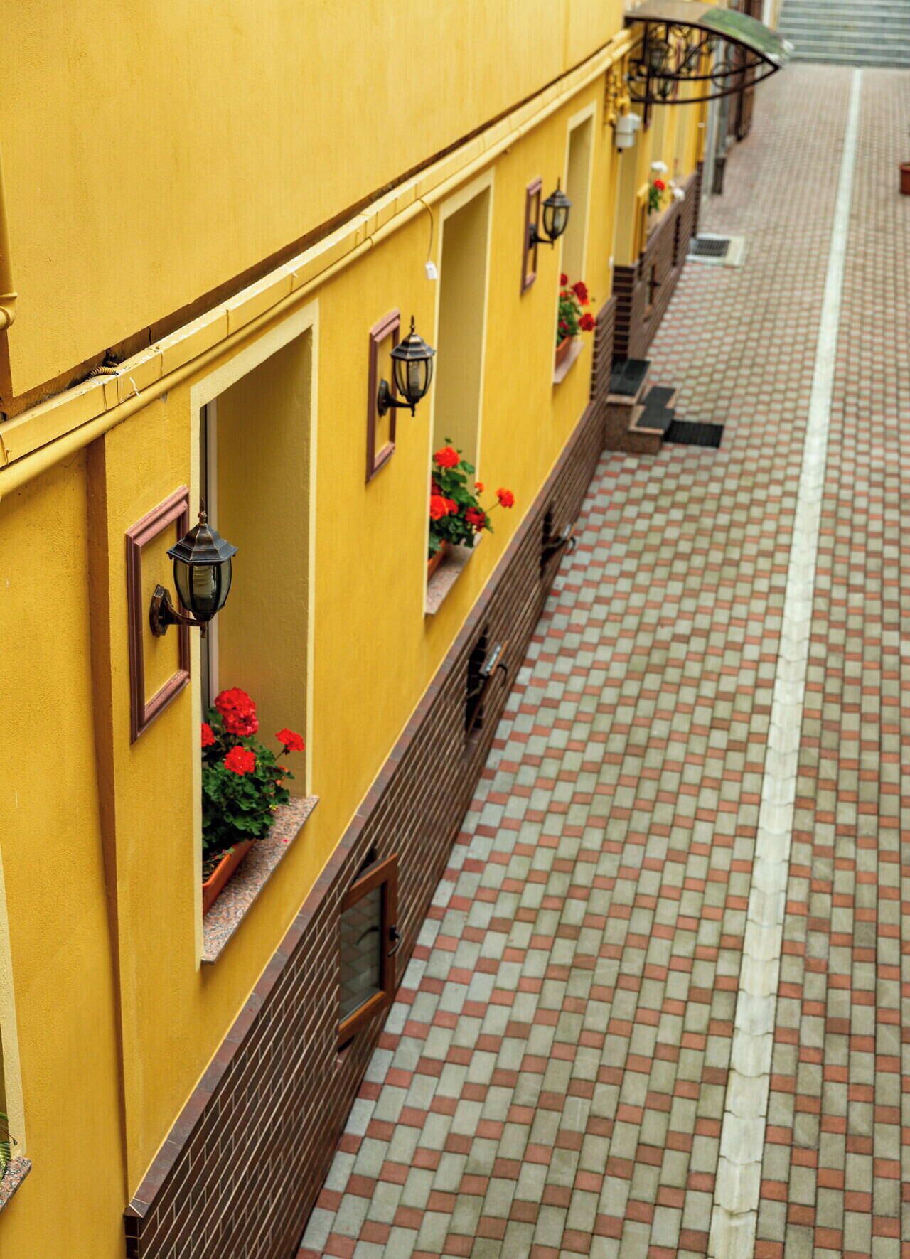 Un edificio amarillo con múltiples ventanas, algunas con jardineras, y un paseo cubierto de baldosas.