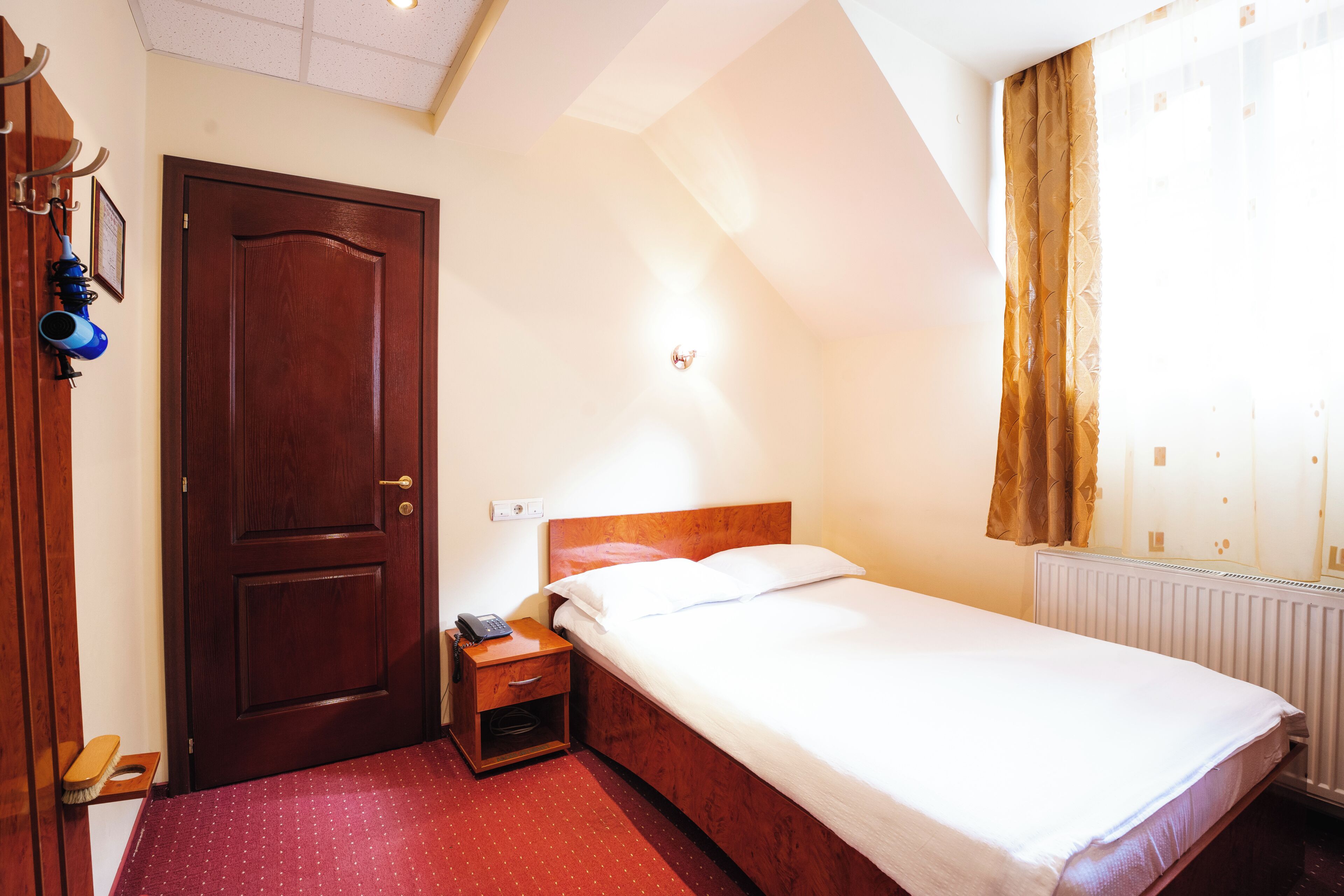 A hotel room with a wooden door, a bedside table with a phone, a bed with white linens, a radiator, and a window with curtains.