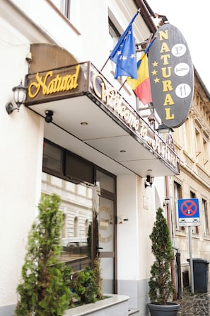 A hotel entrance with a sign reading 'Natura Hotel,' flags, and a no parking sign.
