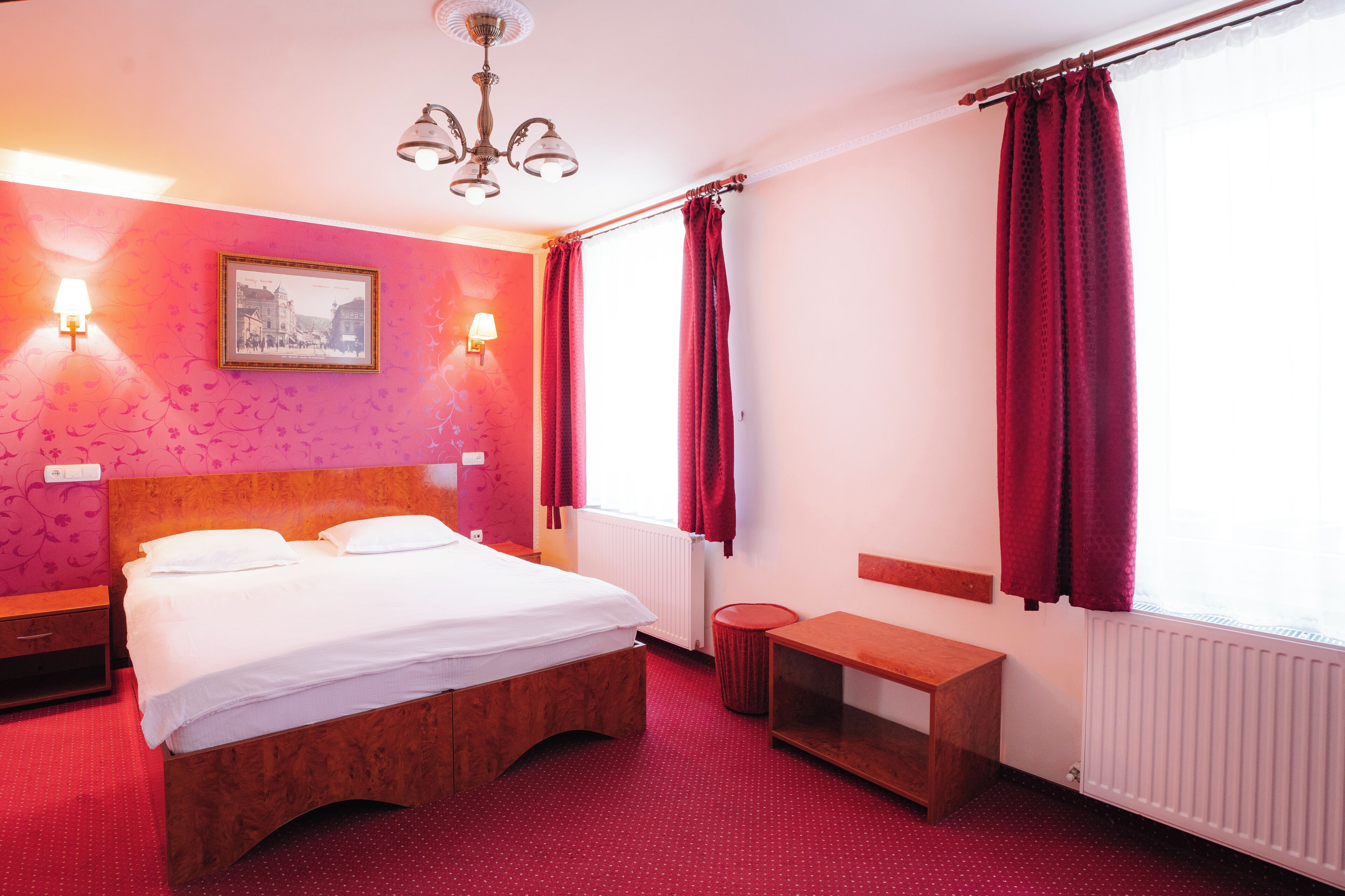 Habitación de hotel con cama, lámparas de noche, un cuadro en la pared, ventana con cortinas y un radiador.