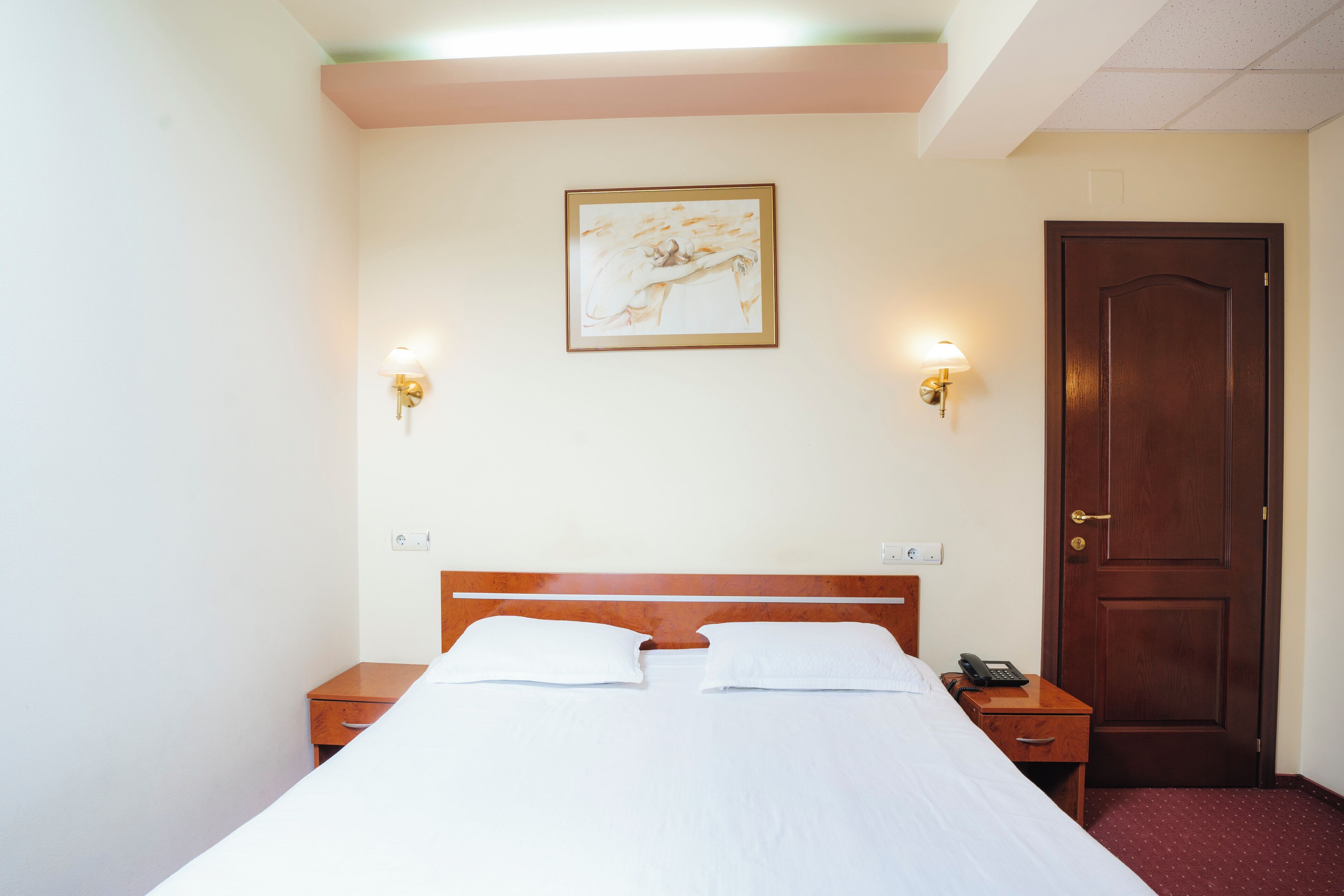 A hotel room with a bed, bedside tables, a wooden door, and wall-mounted light fixtures.