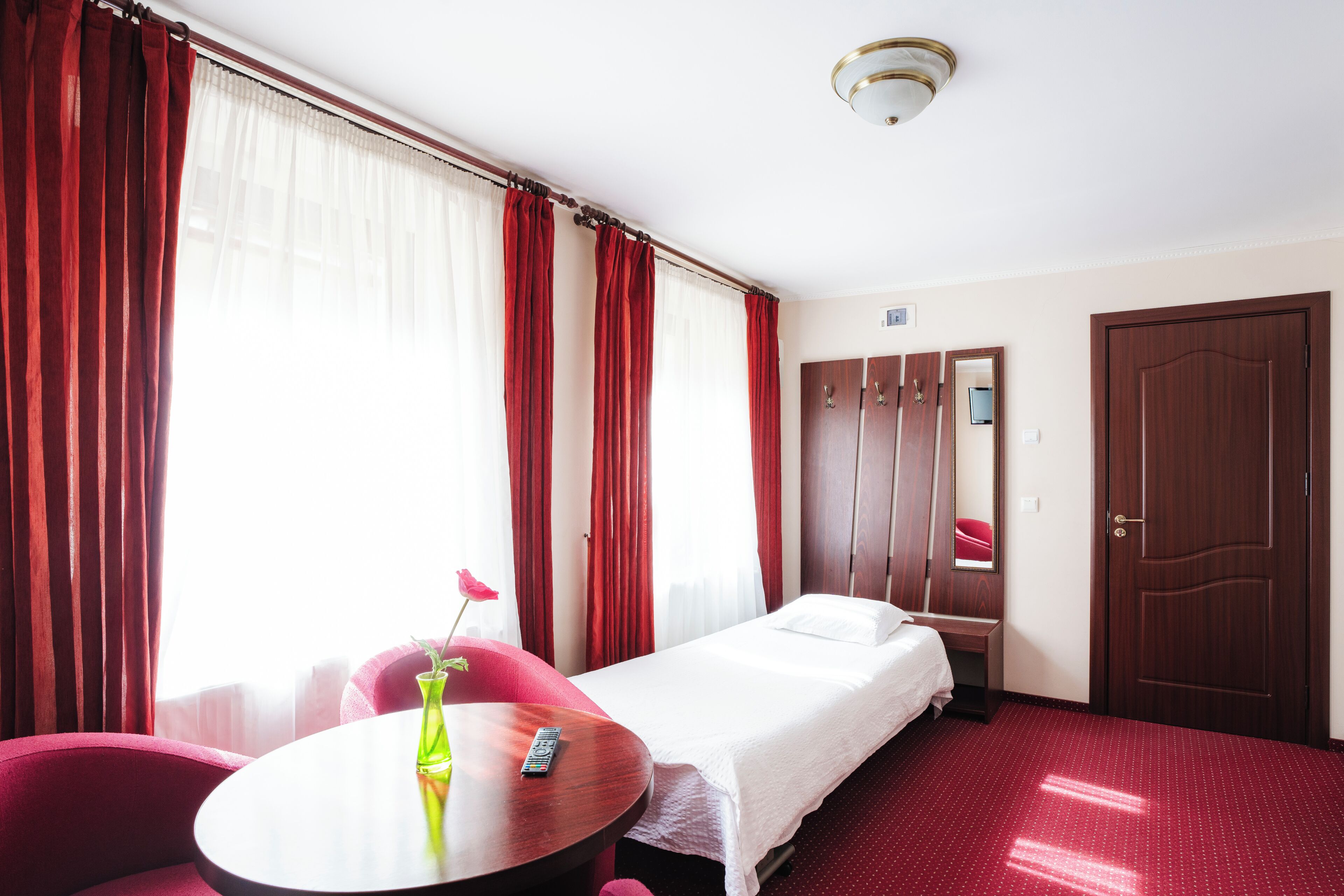 A hotel room with a bed, a small table with a vase and remote, red curtains, a wooden wardrobe, and a door.