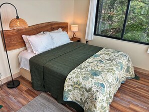 Ein Bett mit Holz-Kopfteil, einer grünen Bettdecke, einem gemusterten Überwurf, einer Nachttischlampe und einem Fenster mit Blick auf einen Wald.