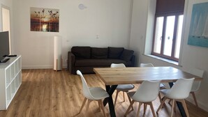 Ett modernt vardagsrum med ett matbord i trä, vita stolar, en brun soffa och en tavla på väggen.