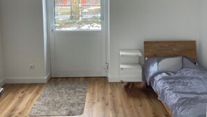 Ett sovrum med trägolv, en säng med ett grått överkast, ett vitt sängbord och ett fönster med utsikt över en röd byggnad.