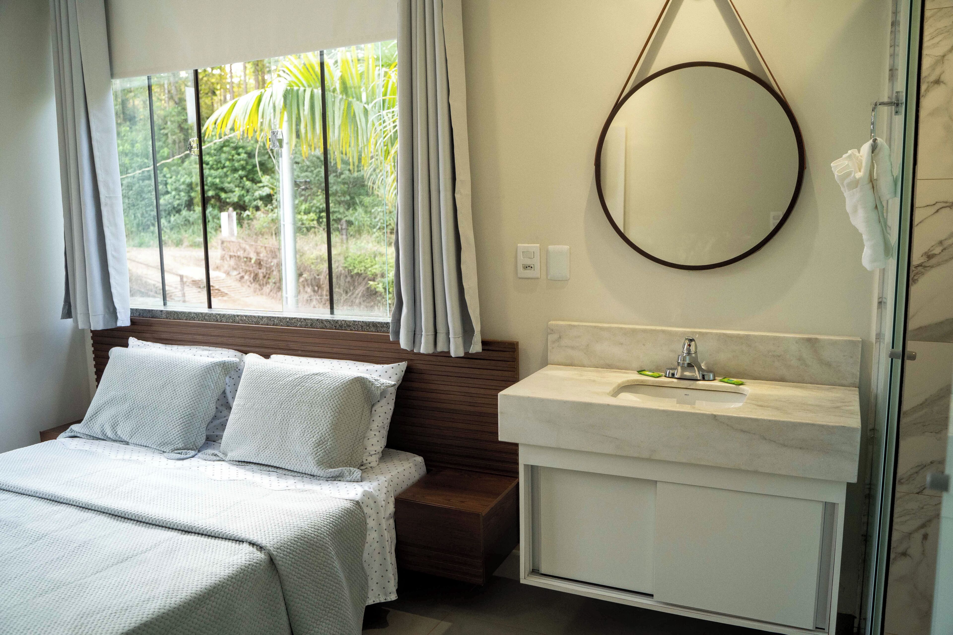 A bedroom with a bed, a bedside table, a window with curtains, and a bathroom with a sink and mirror.