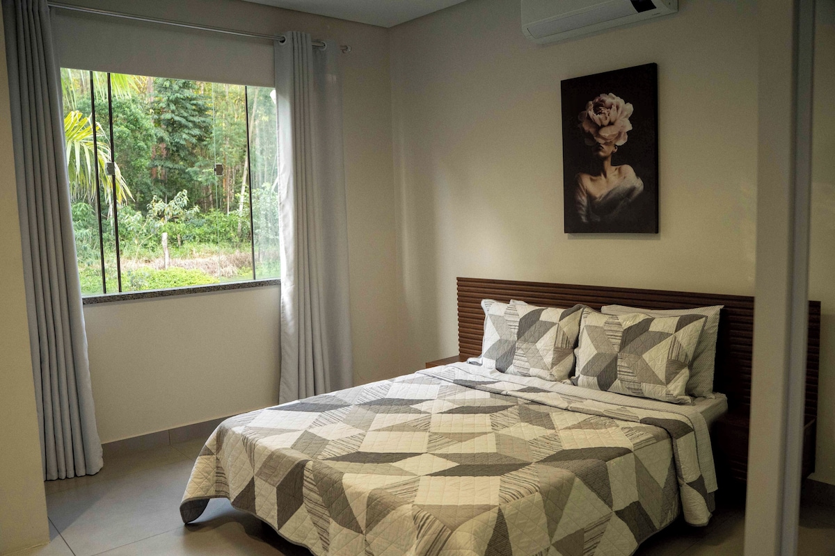 Een slaapkamer met een bed, gordijnen, een schilderij en een raam met uitzicht op groen.