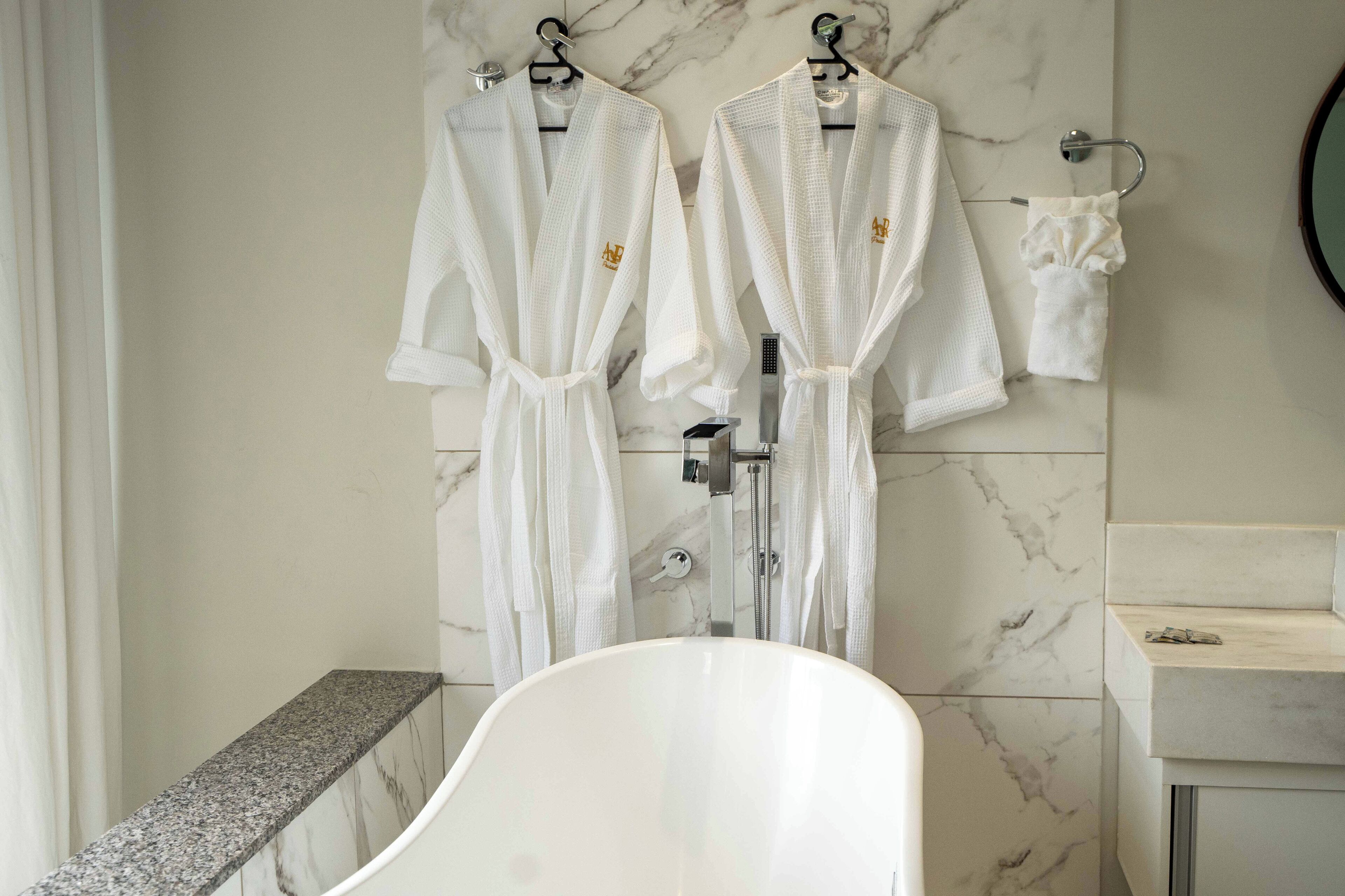 Un baño con dos albornoces blancos colgados en ganchos, una bañera blanca y una pared de mármol.