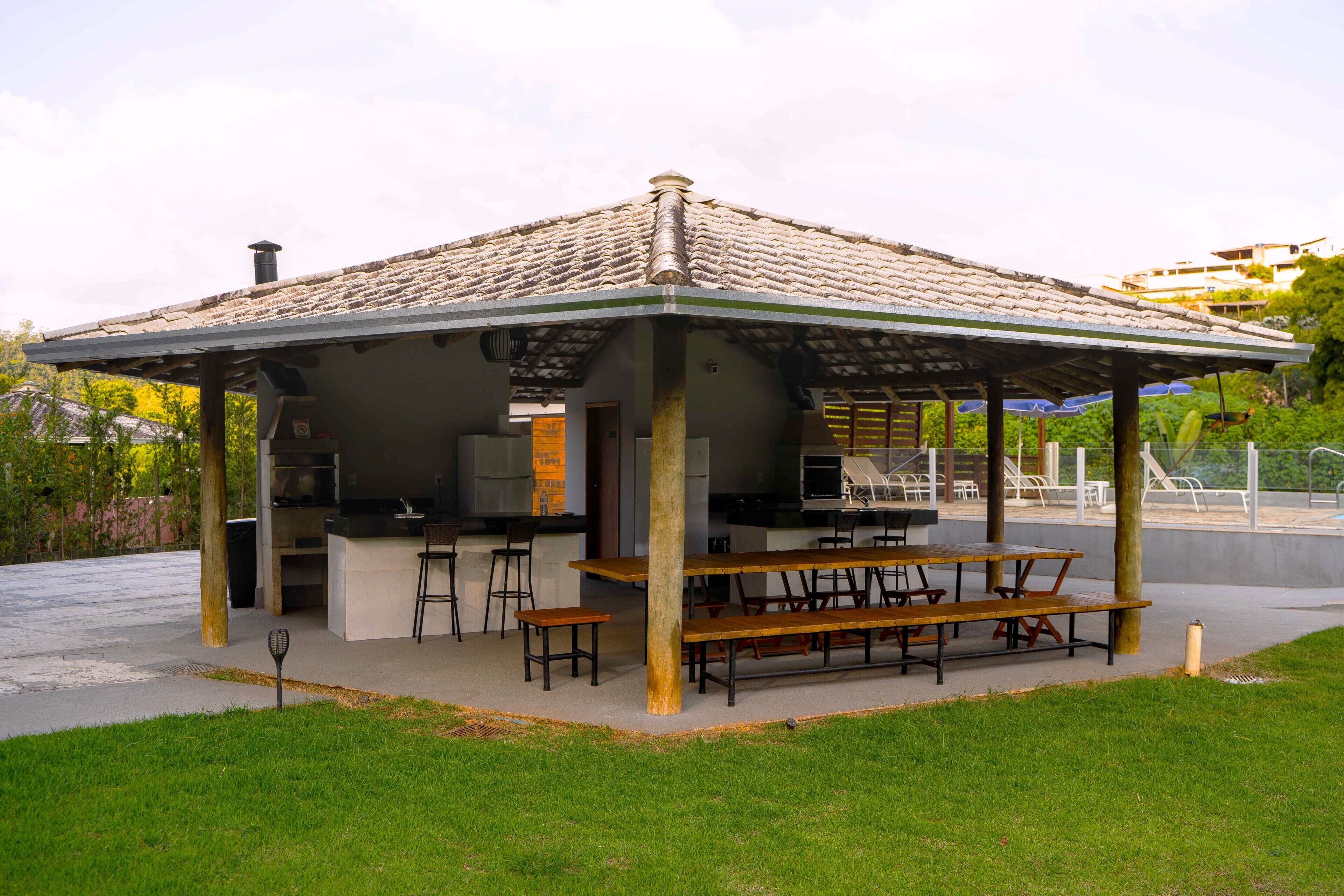 A covered outdoor barbecue area with a grill, seating, and a view of a swimming pool.