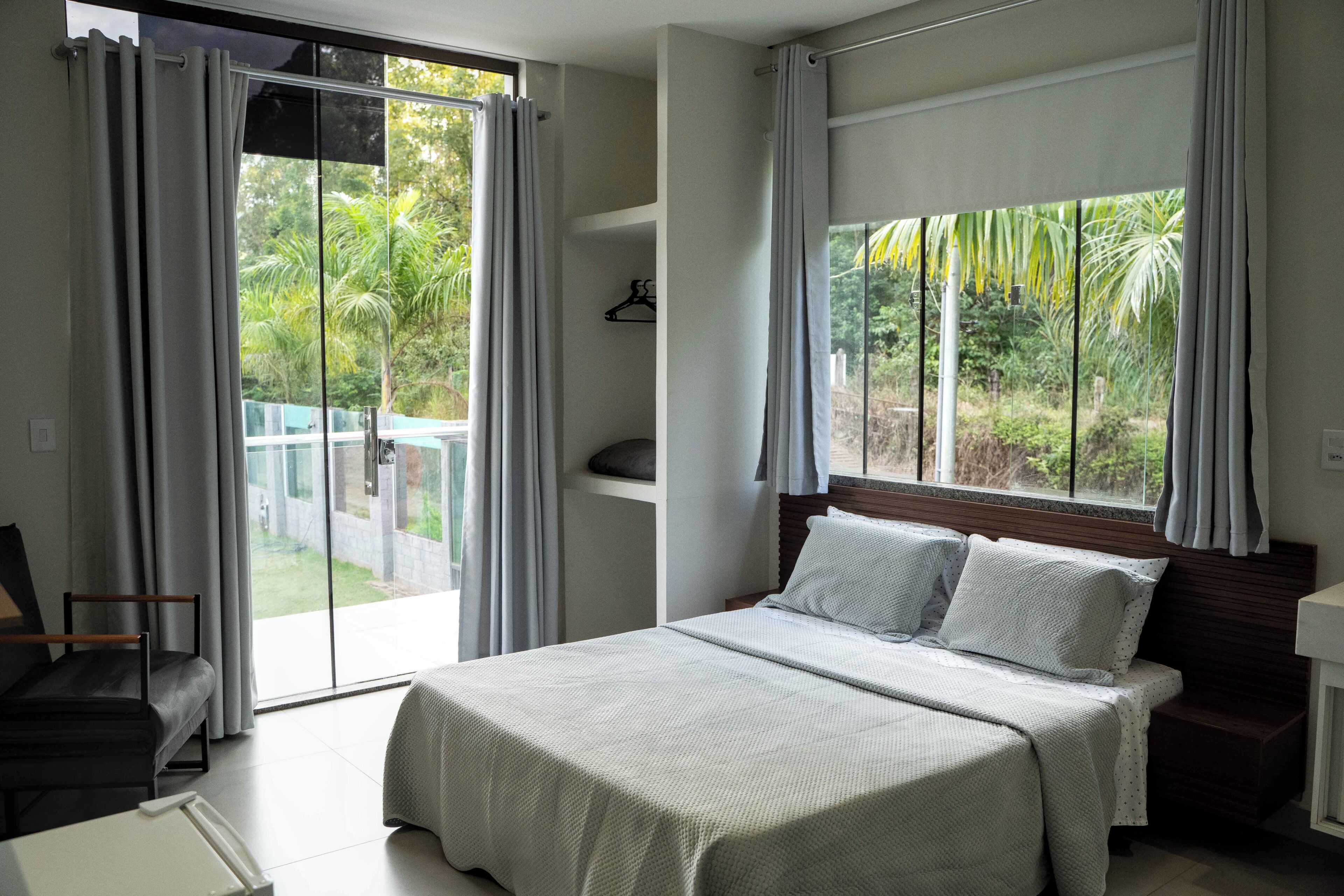 Un dormitorio con una cama grande, una silla, un ventanal con cortinas y vistas a la vegetación.