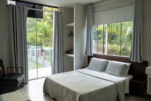 A bedroom with a large bed, a chair, a window with curtains, and a view of greenery outside.