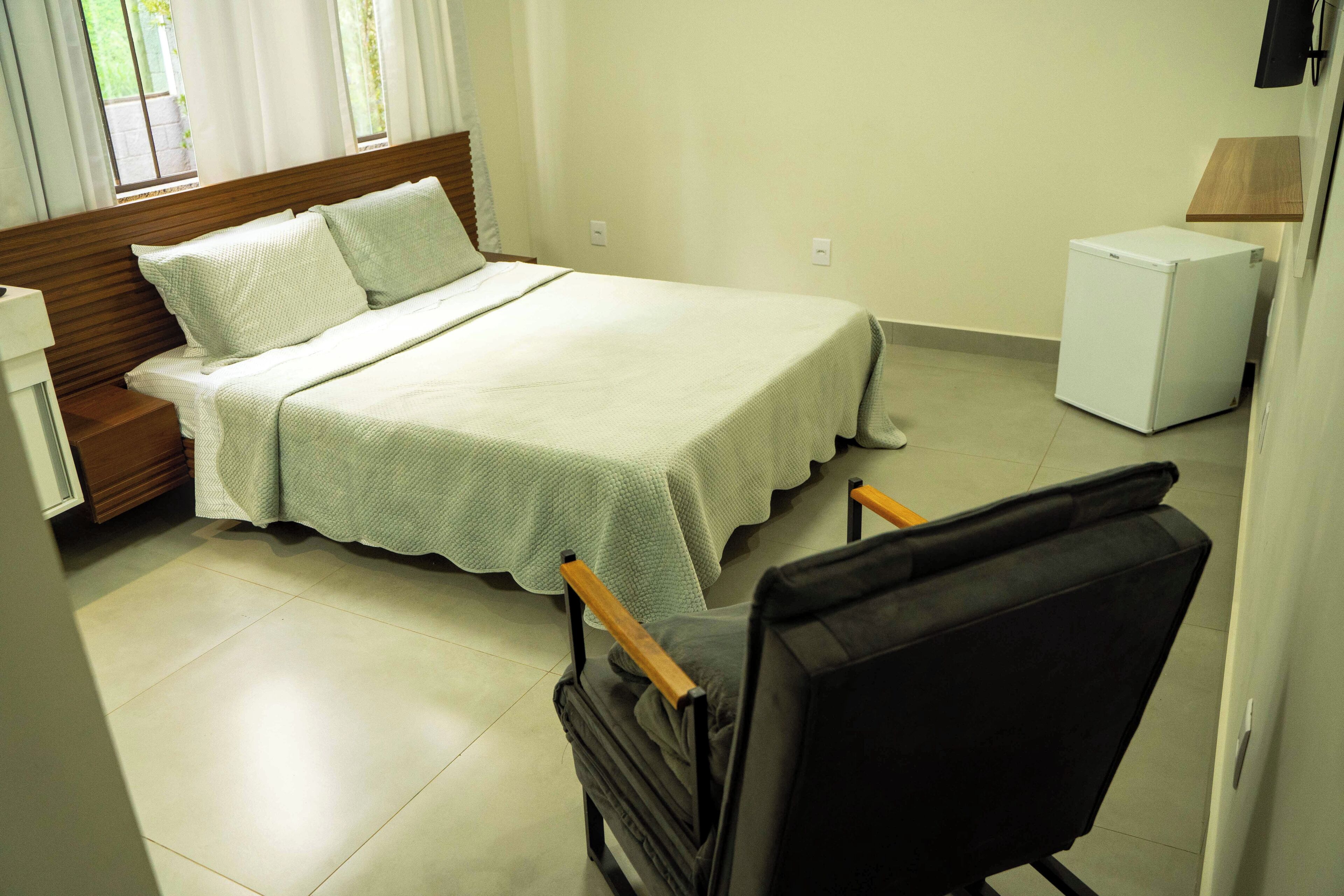 Un dormitorio con cama, una silla, mesita de noche, lámpara y refrigerador.